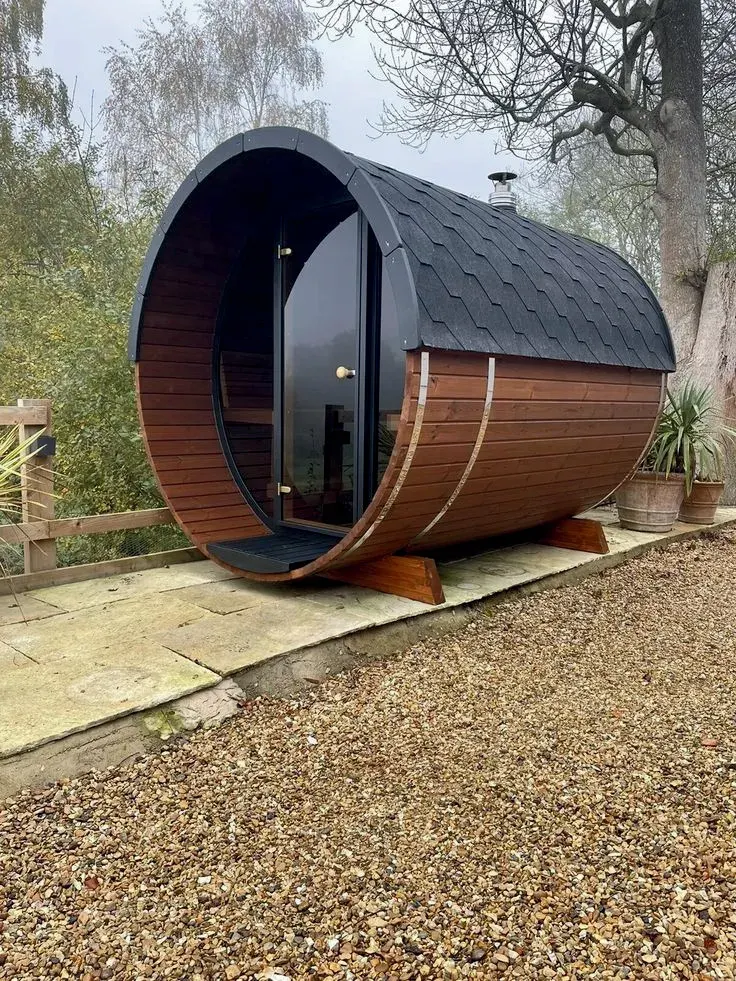 Daş döşənmiş həyətdə kompakt barrel sauna, tünd teqola dam və bacası ilə