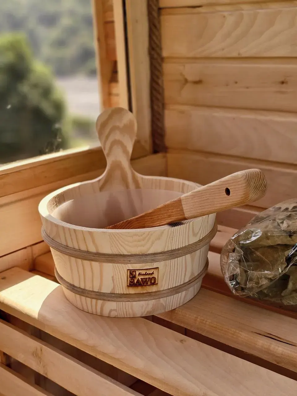 Açıq qapılı taxta sauna, içində təmiz cökə rəflər, qapının yanında yumşaq fırça və təmizləyici ilə vedrə